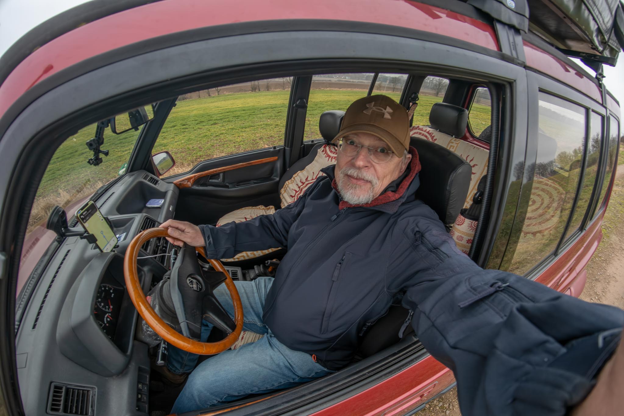 Frank sitzt hinter dem Steuer seines Suzuki Vitara. Er trägt blaue Jeans, eine braune Schirmmütze und eine blaue Jacke. Einen Arm hält er aus dem geöffneten Fenster. Mit einer Action-Cam in der Hand nimmt er ein Selfie auf.