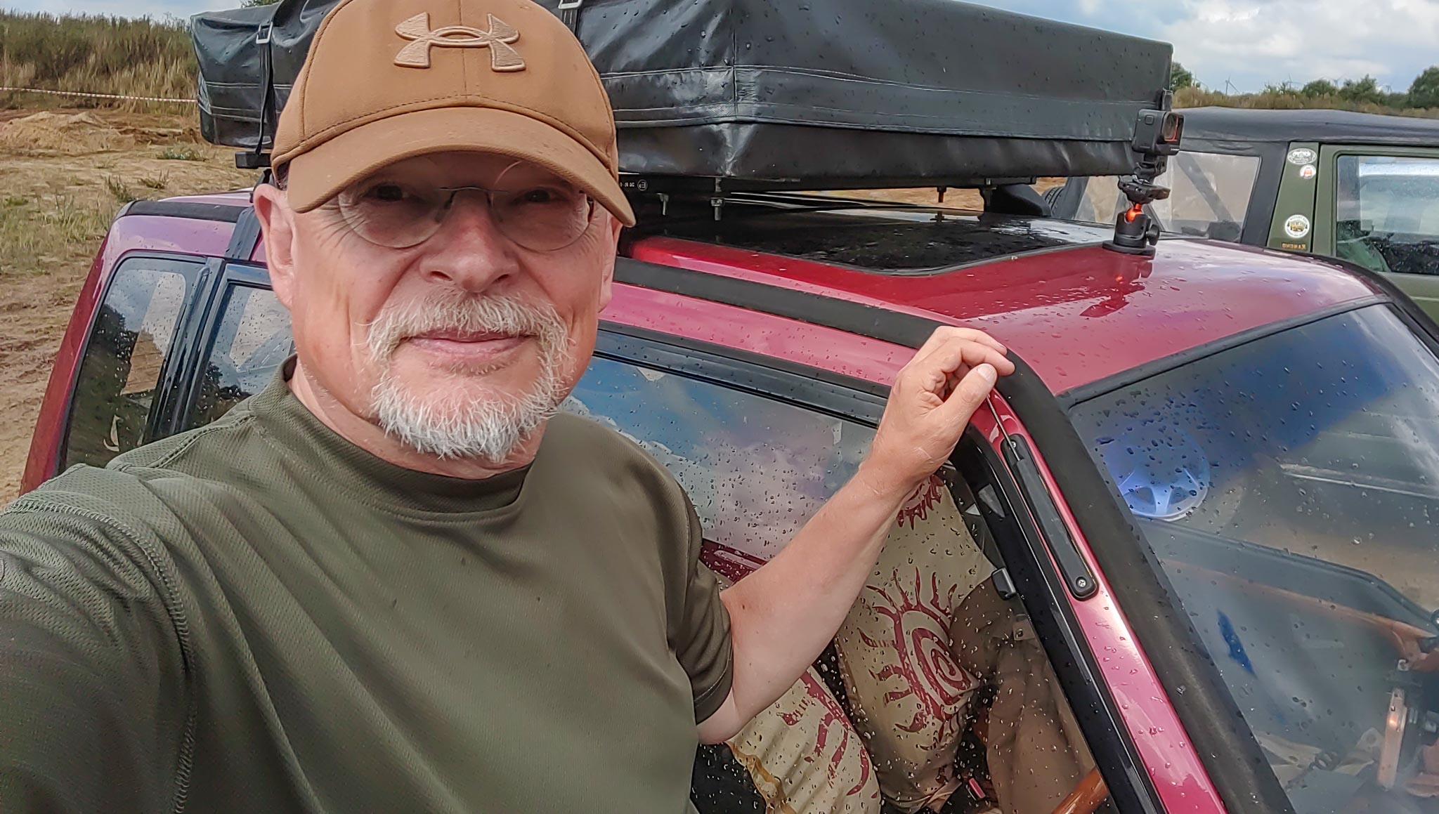 Mann mit Cap und grauem Bart steht neben rotem Suzuki Vitara mit Dachzelt, Selfie im Regen